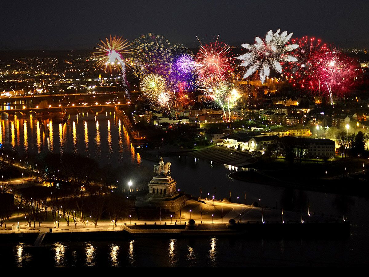 Luftaufnahme von Koblenz bei Nacht mit bunten Feuerwerken über dem Deutschen Eck, Flüssen Rhein und Mosel, beleuchteten Brücken und Stadtlichtern.