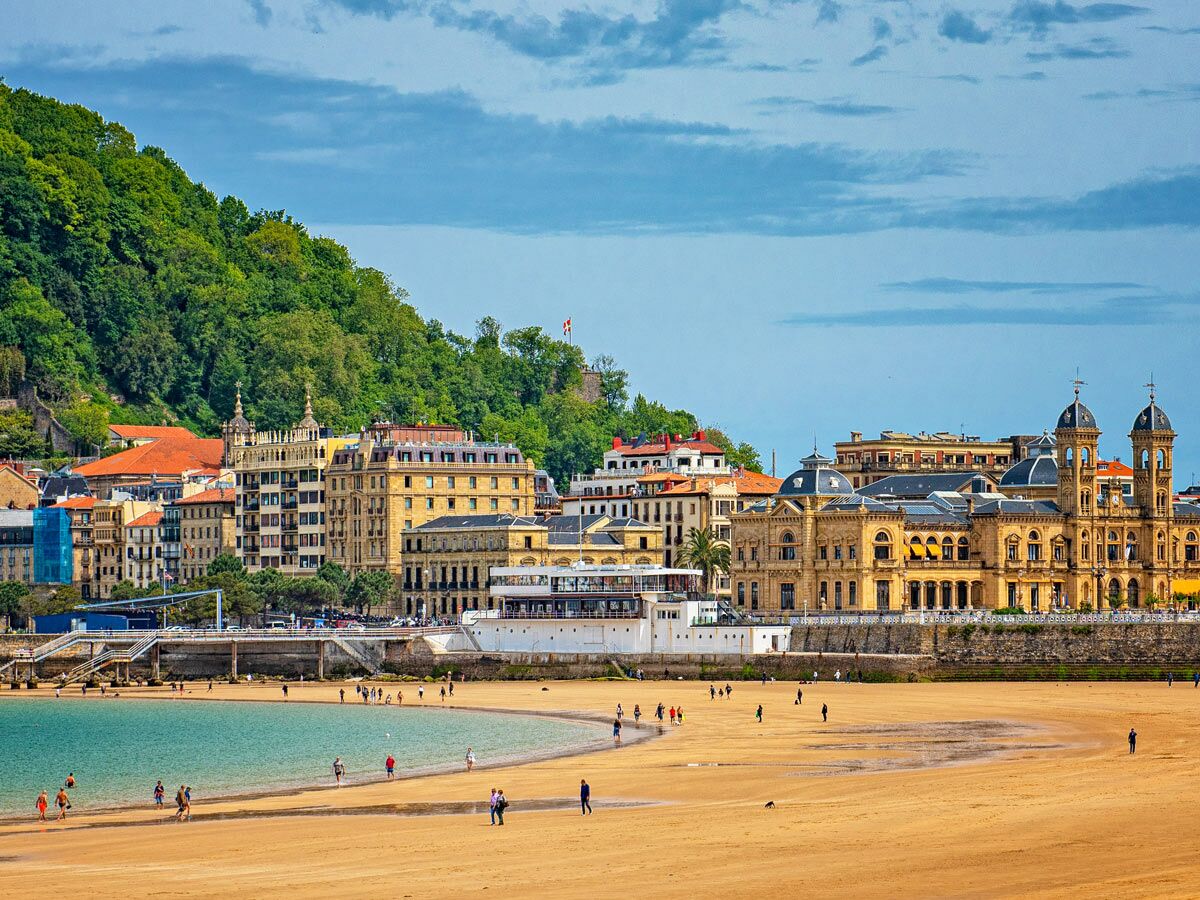 Panoramablick auf San Sebastián mit goldenem La Concha Strand, türkisfarbenem Meer und historischen Gebäuden am Fuß grüner Hügel.