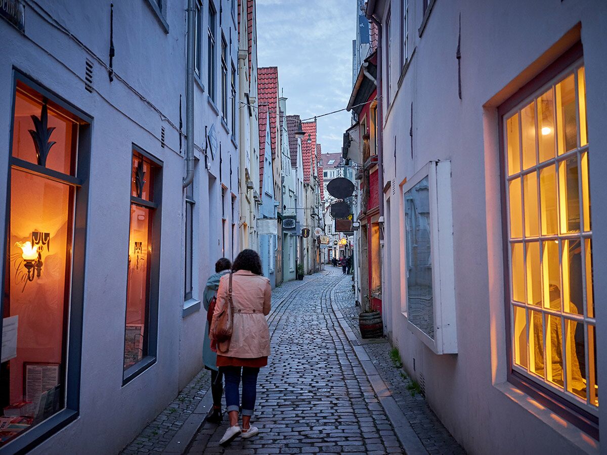 Zwei Personen gehen durch eine schmale, beleuchtete Kopfsteinpflastergasse in einer historischen Altstadt bei Abenddämmerung.