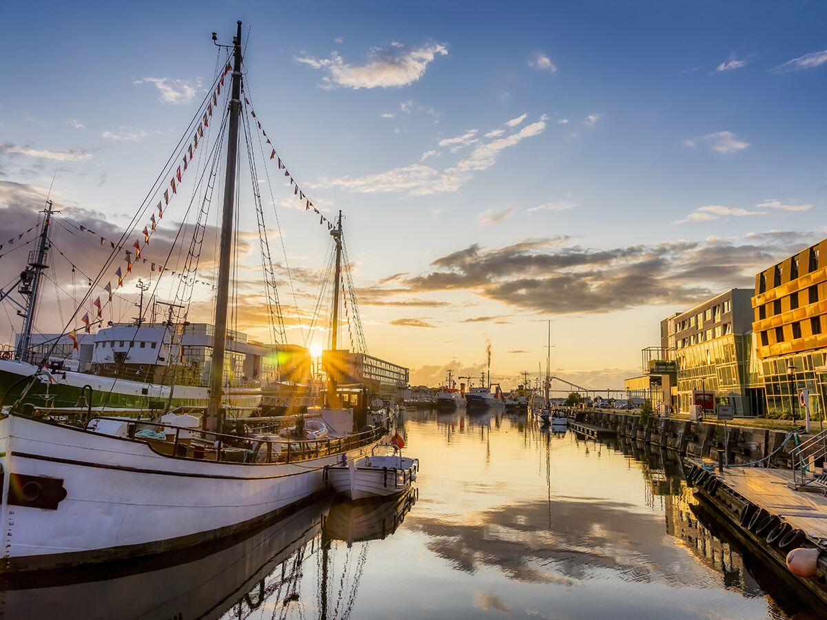 Goldener Sonnenuntergang in einem Hafen mit Segelschiffen, Booten und modernen Gebäuden, die sich im ruhigen Wasser spiegeln.