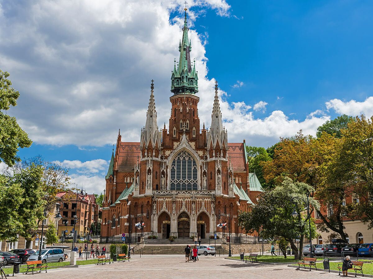 Die reich verzierte neugotische St. Josefskirche in Krakau, Polen, mit hohem Turm, Ziegelfassade und Menschen auf dem sonnigen Platz.