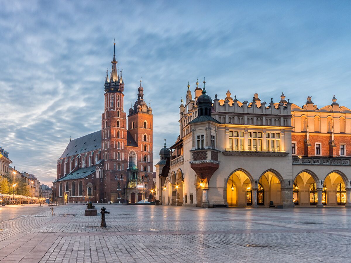 Historischer Hauptmarkt in Krakau mit der beleuchteten Marienkirche und Tuchhallen bei Dämmerung unter bewölktem Himmel.
