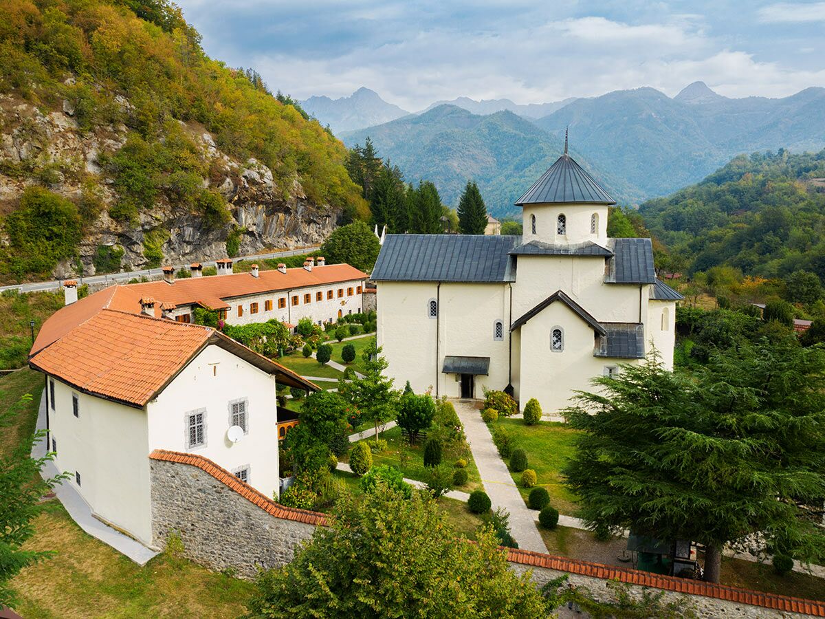 Weißes orthodoxes Kloster mit dunklem Dach, umgeben von grünen Bergen und bewaldeten Hügeln.