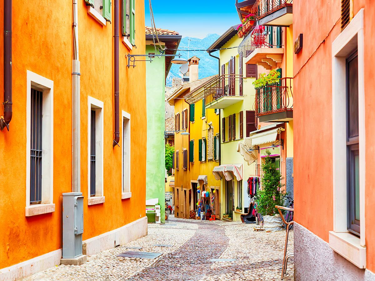 Enge, bunte Gasse mit orangefarbenen, gelben und grünen Häusern, Balkonen und Kopfsteinpflaster. Blauer Himmel und Berge im Hintergrund.