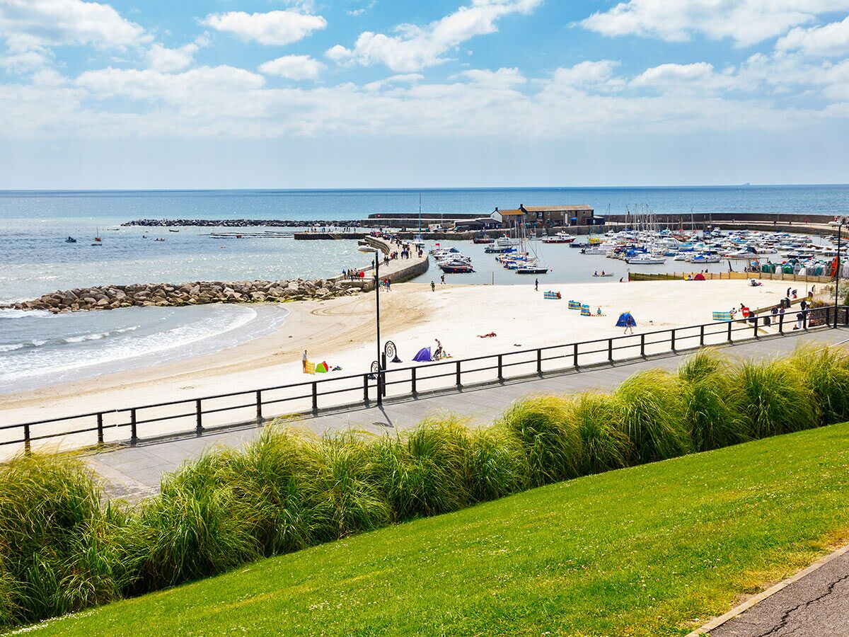 Panoramablick auf einen belebten Hafen mit vielen Booten, Sandstrand, Wellenbrechern und Spaziergängern an einem sonnigen Küstentag.