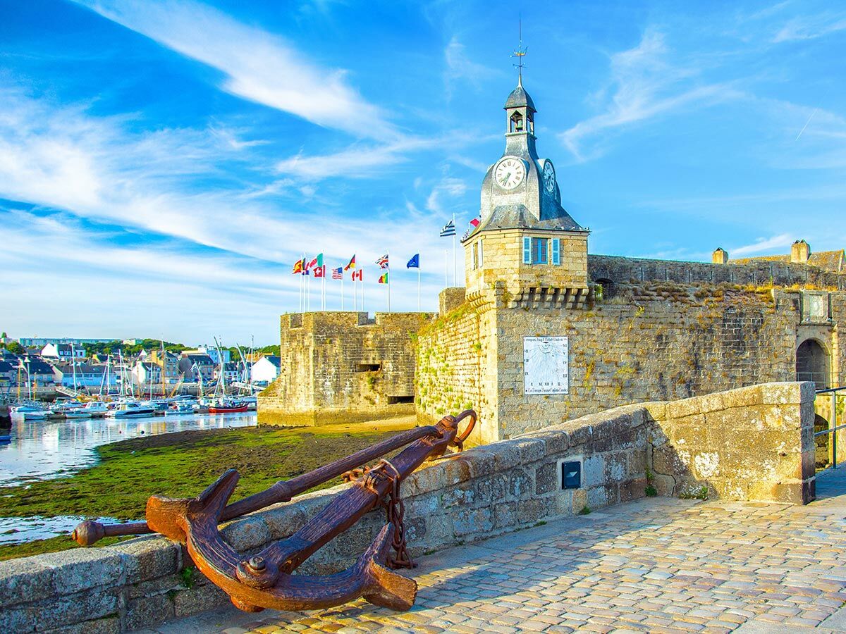 Historische Steinfestung mit Uhrturm, Sonnenuhr und rostigem Anker über einem geschäftigen Hafen mit Booten unter blauem Himmel.
