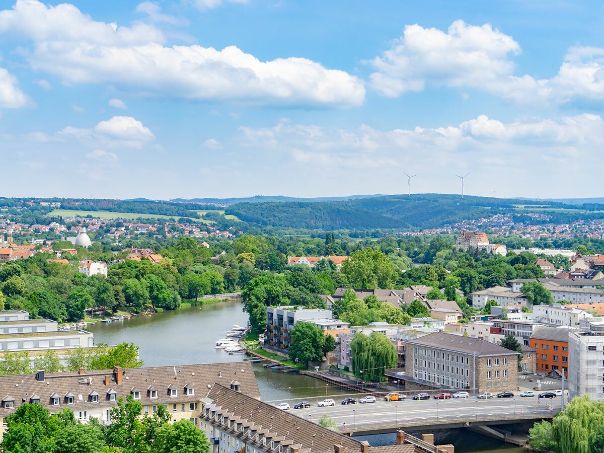 Luftbild einer grünen Stadt mit Fluss, Booten, Brücke, Gebäuden, einem Dom und Windrädern unter blauem Himmel.