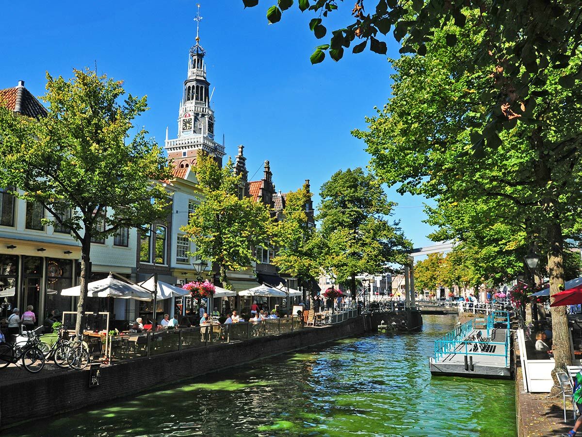 Ansicht einer historischen Grachtenstadt mit markantem Kirchturm, belebten Cafés am Kanal, grünen Bäumen und Fahrrädern.