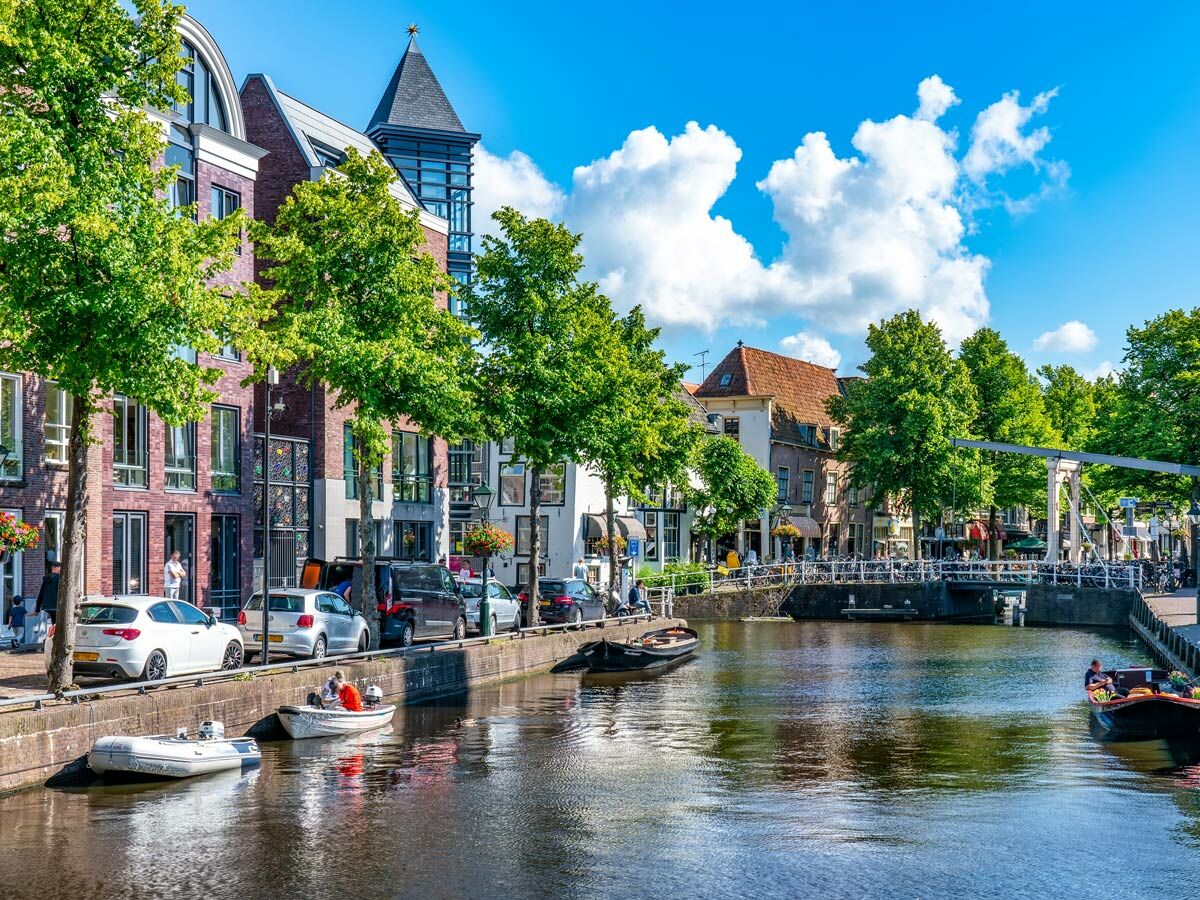 Sonniger Kanal in Leiden, Niederlande, mit historischen und modernen Gebäuden, grünen Bäumen, Booten und einer Zugbrücke.