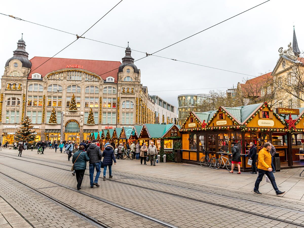 Belebter Weihnachtsmarkt in Erfurt am Anger mit geschmückten Holzbuden, Karstadt-Gebäude und Passanten unter winterlichem Himmel.