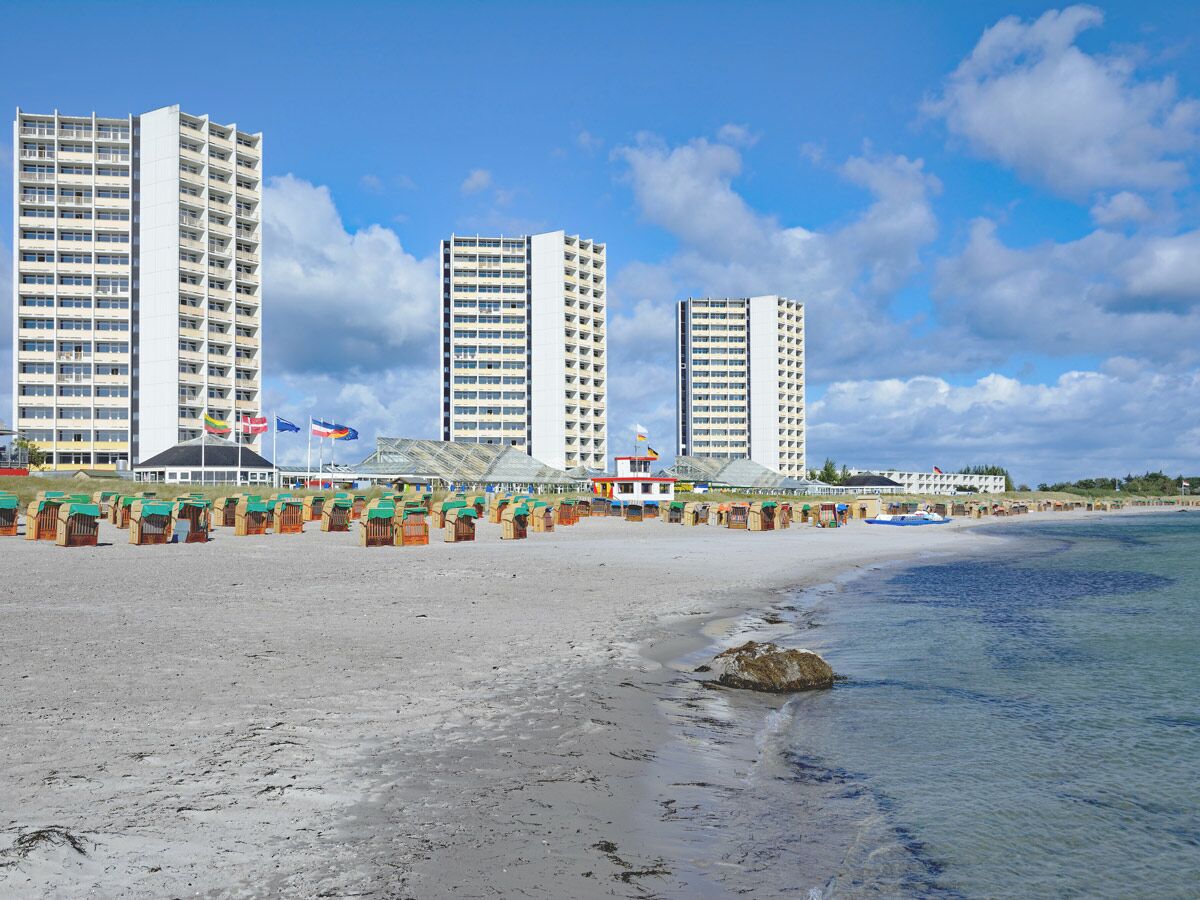 Weiter Sandstrand mit vielen bunten Strandkörben vor modernen Hochhäusern unter blauem Himmel und weißen Wolken.