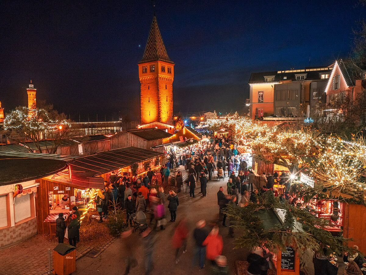 Belebter Weihnachtsmarkt bei Nacht, viele Menschen flanieren zwischen hell beleuchteten Ständen und einem historischen Turm.