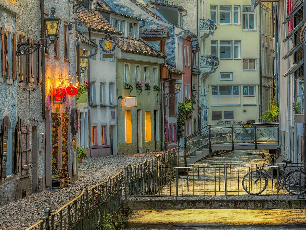Malerische historische Gasse mit Kopfsteinpflaster, Kanal und Brücken in warmem Abendlicht. Bunte Häuser, Laternen und ein Fahrrad.