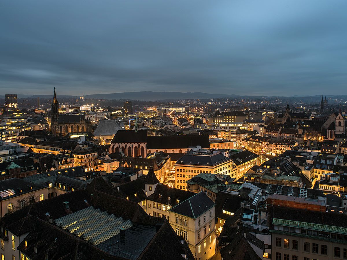 Panoramablick auf das beleuchtete Basel bei Abenddämmerung mit historischen Kirchen, modernen Gebäuden und Stadtlichtern vor Bergen.