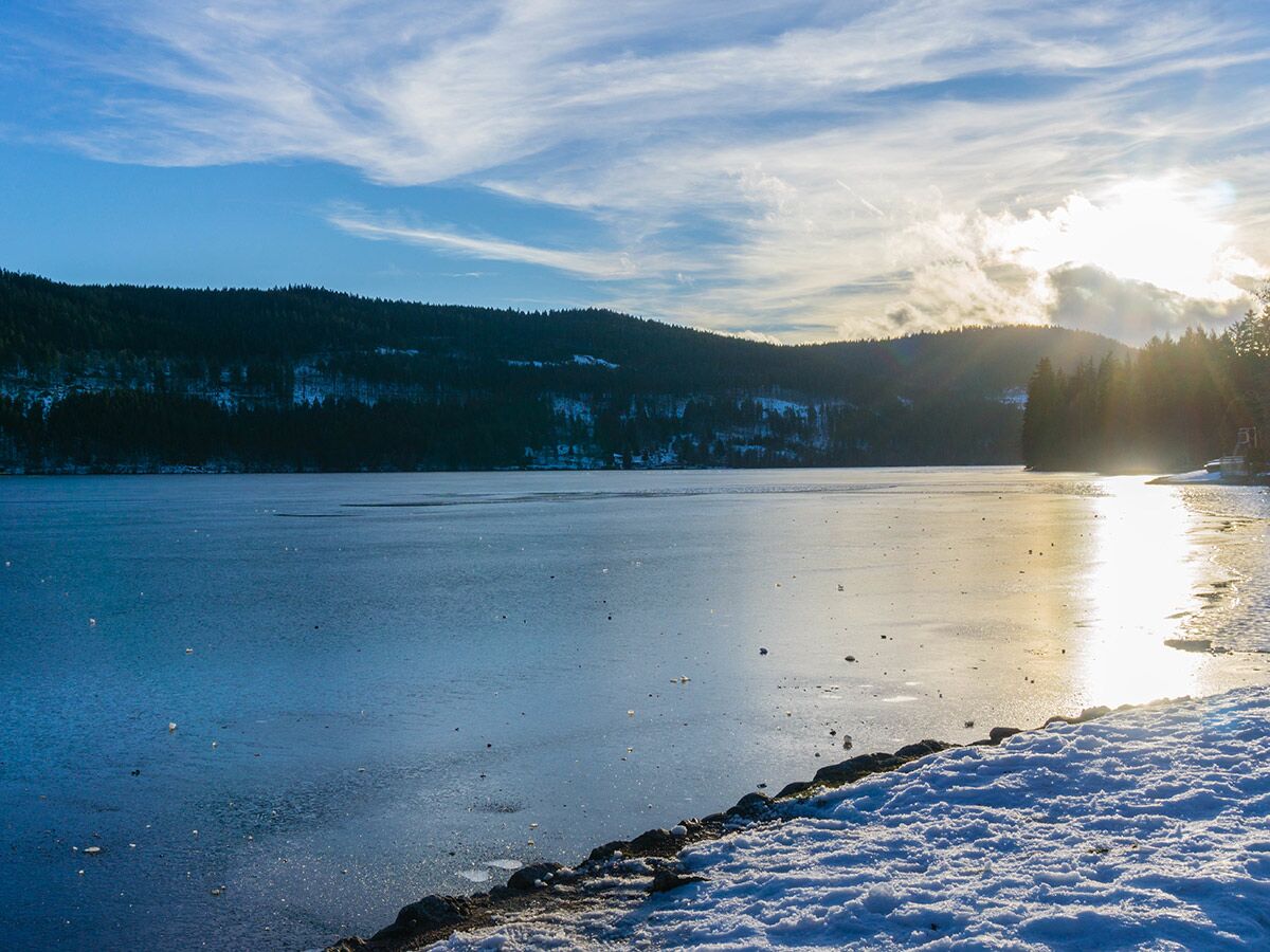 Malerische Winterlandschaft mit teilweise gefrorenem See, schneebedeckten Waldhügeln und heller Sonne, die sich im Eiswasser spiegelt.