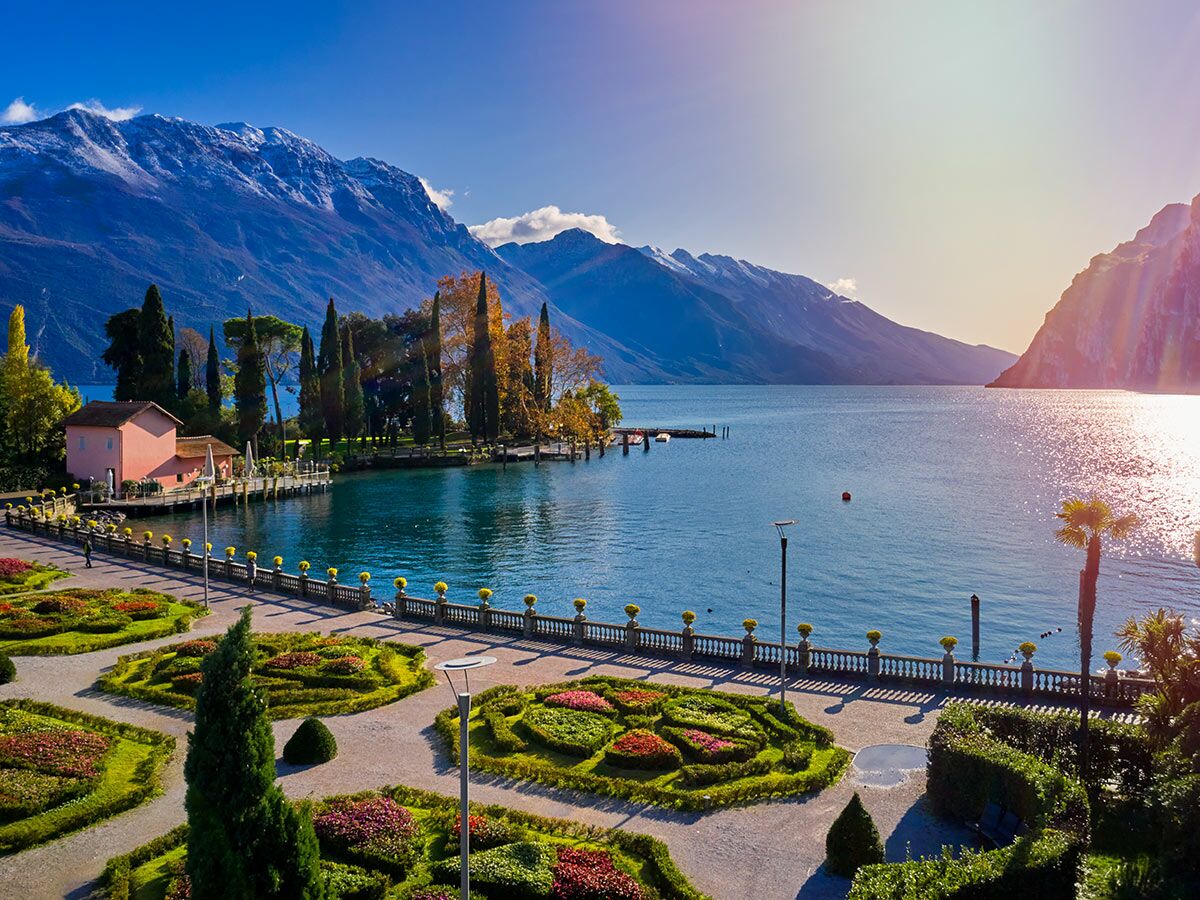 Panoramablick auf den Gardasee mit kunstvollen Gärten, einer Uferpromenade, einem rosa Haus und schneebedeckten Bergen im Herbst bei Sonnenschein.