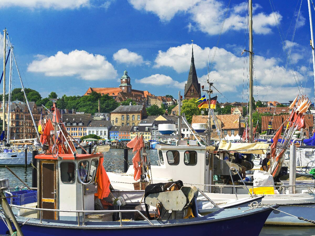 Hafen Flensburg mit Fischer- und Segelbooten, im Hintergrund historische Stadtgebäude, Kirchen und der blaue Himmel mit Wolken.