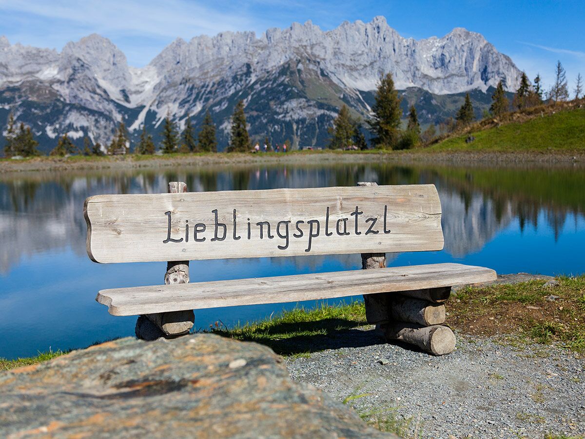 Holzbank "Lieblingsplatzl" am Bergsee mit Alpenpanorama bei Sonnenschein. Idyllischer Ort zur Entspannung.