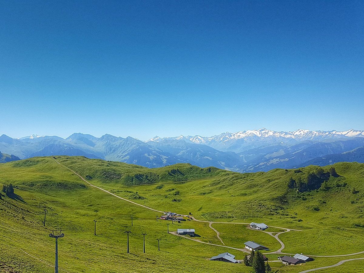 Panoramablick auf eine sonnige Alpenlandschaft mit grünen Wiesen, vereinzelten Hütten und schneebedeckten Bergen am Horizont.