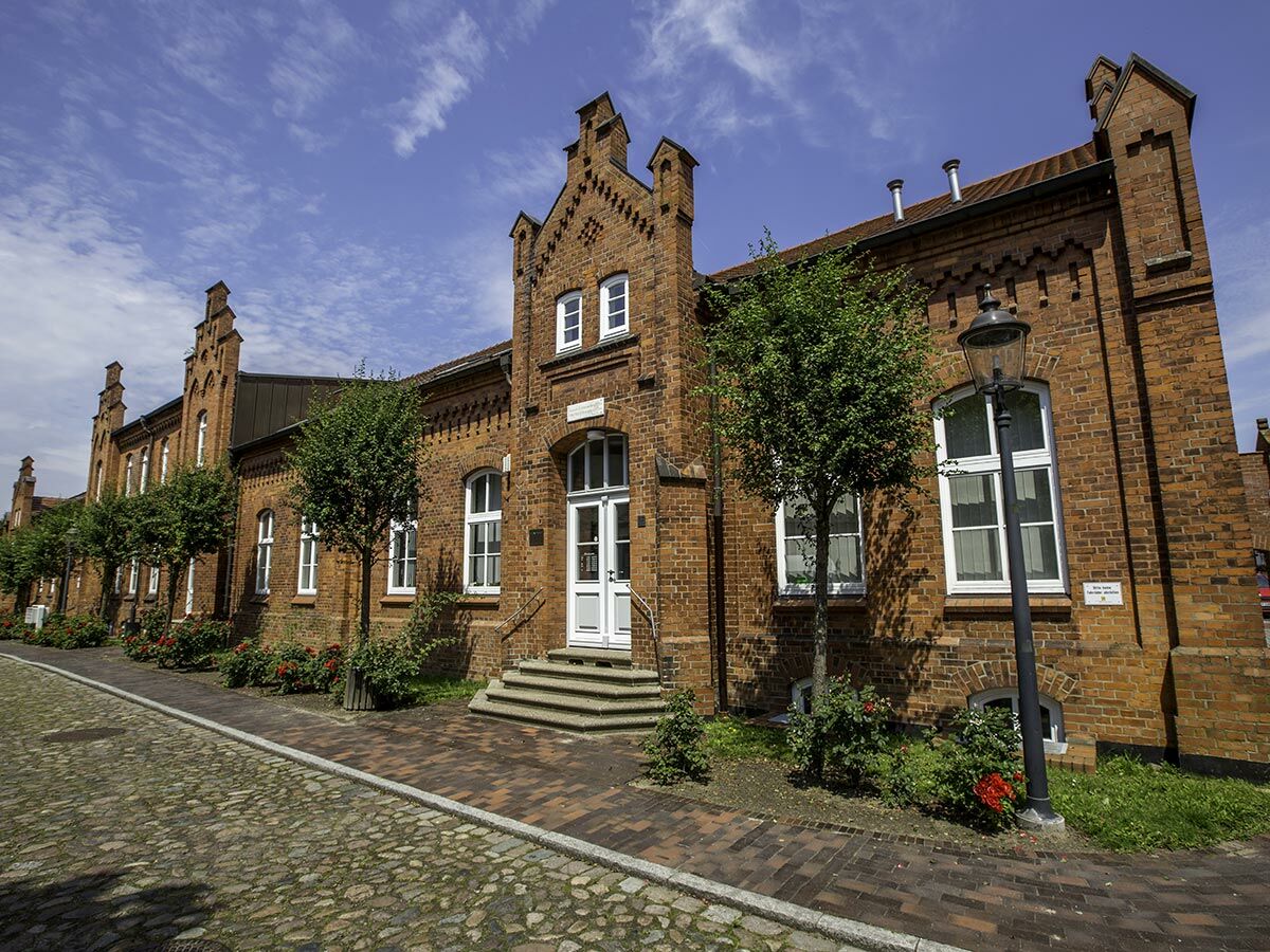 Alte Backsteinarchitektur mit markanten Giebeln, weißen Fenstern, Bäumen und Kopfsteinpflasterstraße unter blauem Himmel.