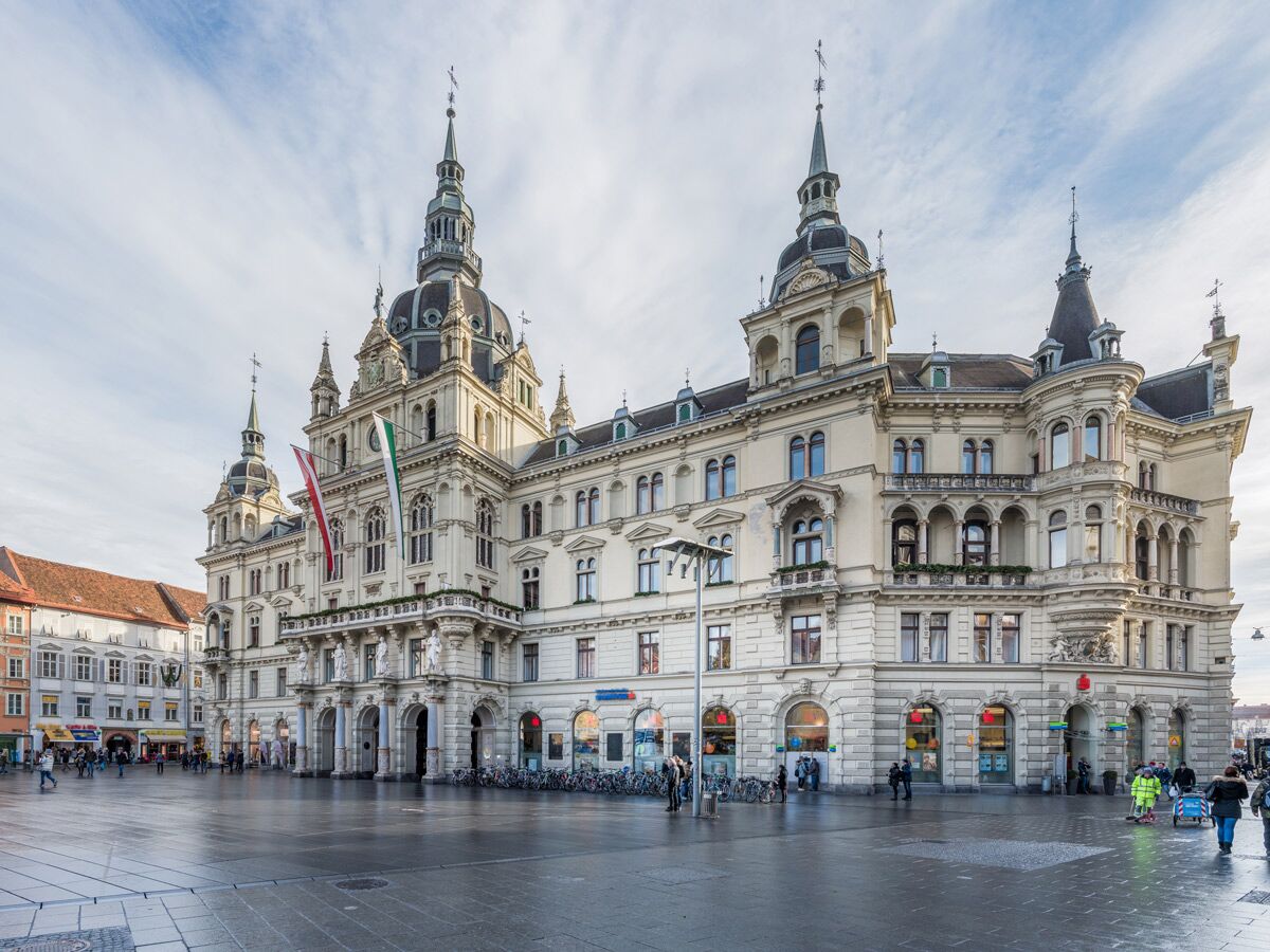 Ansicht des Grazer Rathauses mit seinen Türmen und der österreichischen Flagge am belebten Hauptplatz bei Tag.