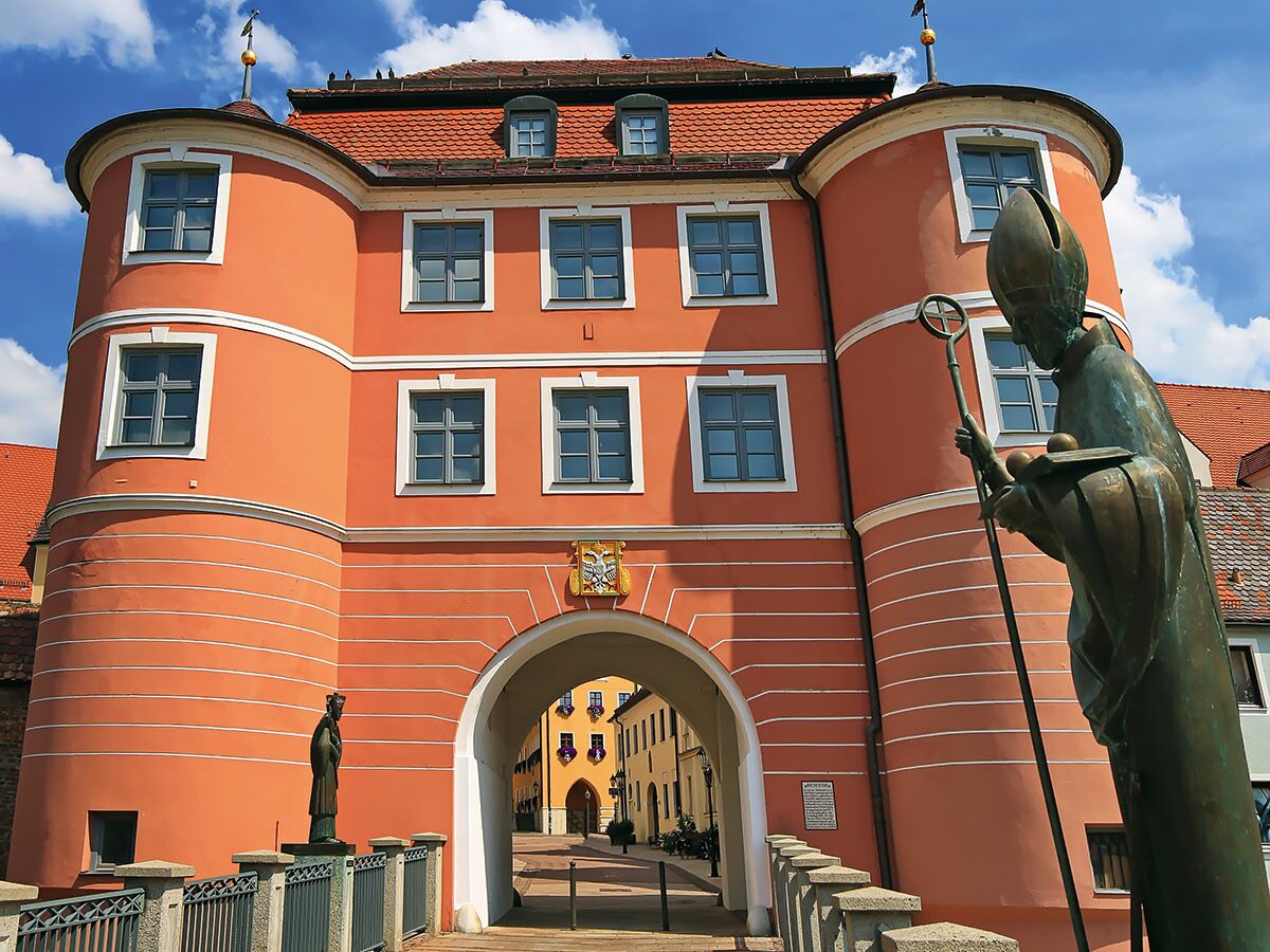 Historisches Himmelsreich Torhaus Merseburg mit korallenfarbener Fassade, Bogen und Bischofsstatue vor blauem Himmel.