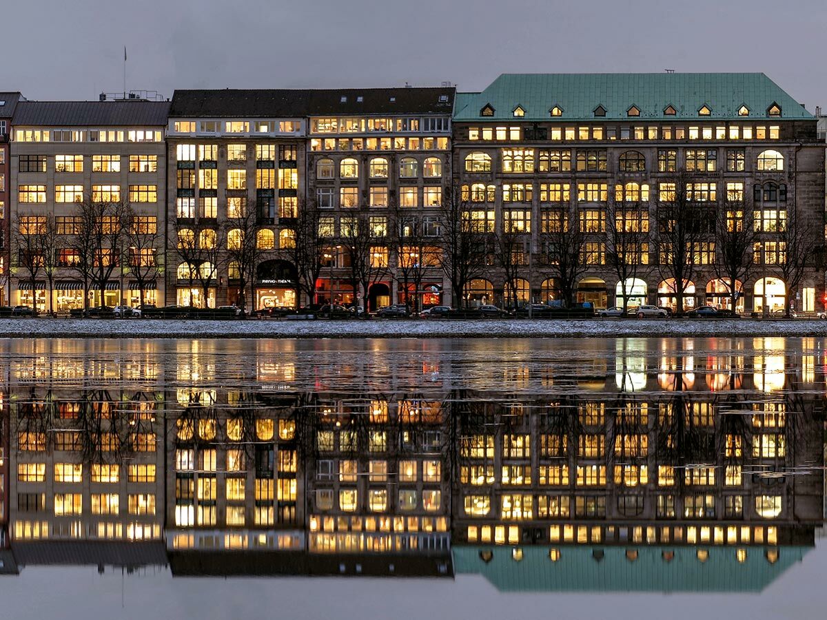 Beleuchtete historische Gebäude spiegeln sich in leicht zugefrorenem Wasser bei Dämmerung, warme Lichter in den Fenstern sichtbar.