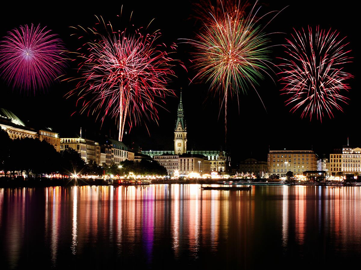 Spektakuläres Feuerwerk in verschiedenen Farben über dem Hamburger Rathaus bei Nacht, gespiegelt im Wasser.