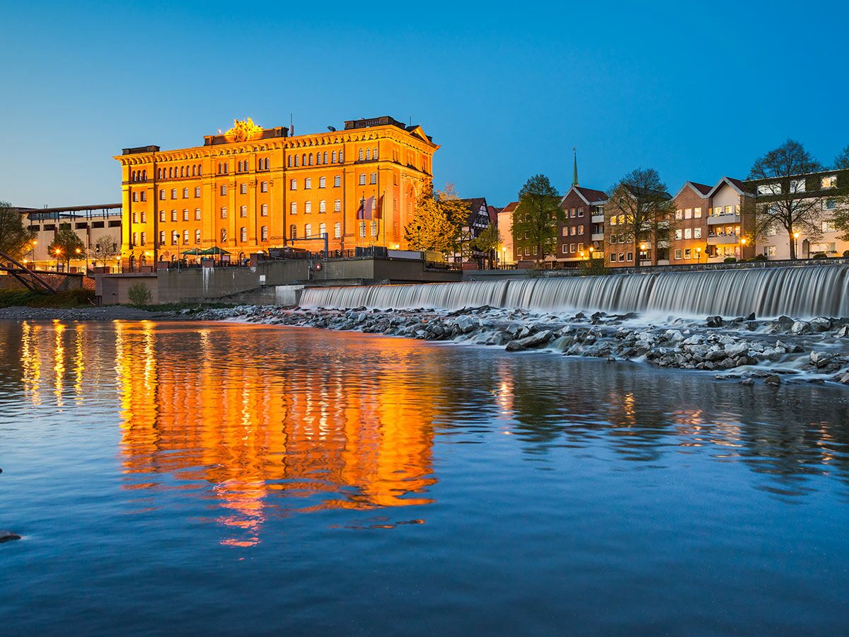 Abendliche Stadtansicht mit beleuchtetem historischen Gebäude und Häusern am Fluss, Wasserfall und starken Reflexionen im blauen Wasser.