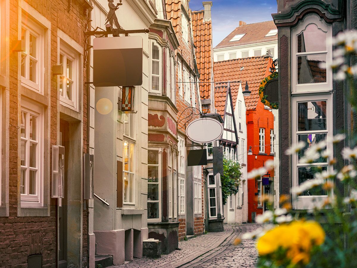 Malerische Altstadtgasse mit Kopfsteinpflaster und historischen Gebäuden, darunter Fachwerkhäuser. Leere Ladenschilder und bunte Blumen im unscharfen Vordergrund.