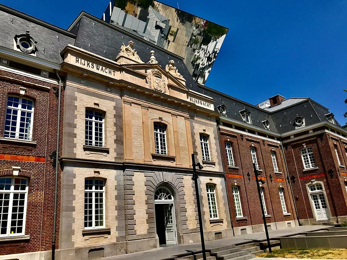 Historisches Rijkswachtgebäude aus Ziegel und Stein, beschriftet mit "RIJKSWACHT", mit moderner spiegelnder Architektur im Hintergrund unter blauem Himmel.