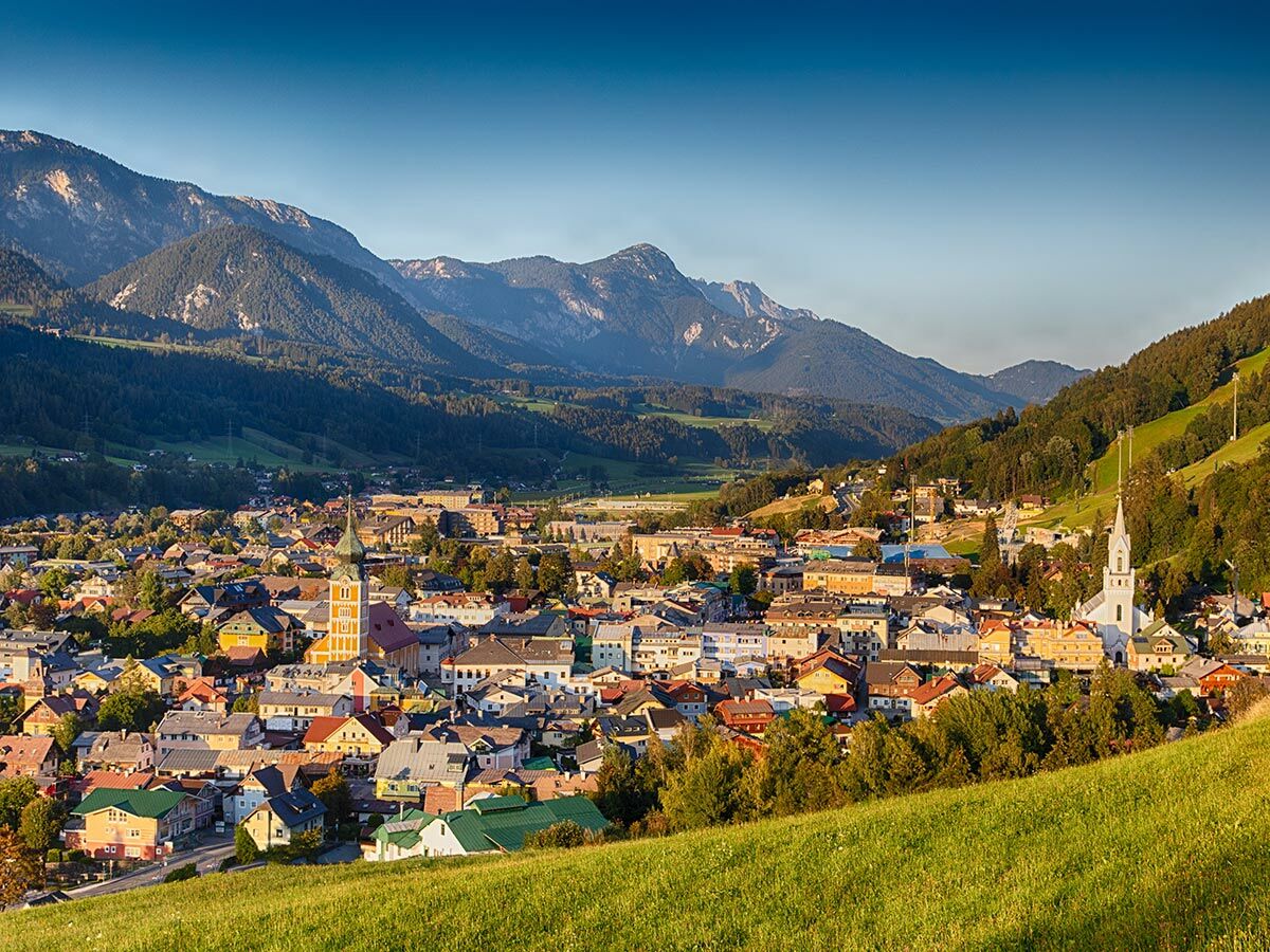 Panorama einer malerischen Bergstadt im Tal, umgeben von grünen Hügeln und majestätischen Bergen mit bunten Häusern und Kirchen.