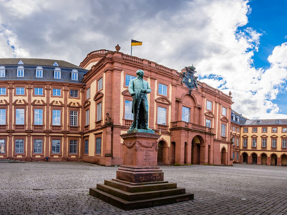 Bronzestatue von Schiller vor dem Mannheimer Schloss, einem großen Barockgebäude mit gelb-roter Fassade und Kopfsteinpflasterhof.