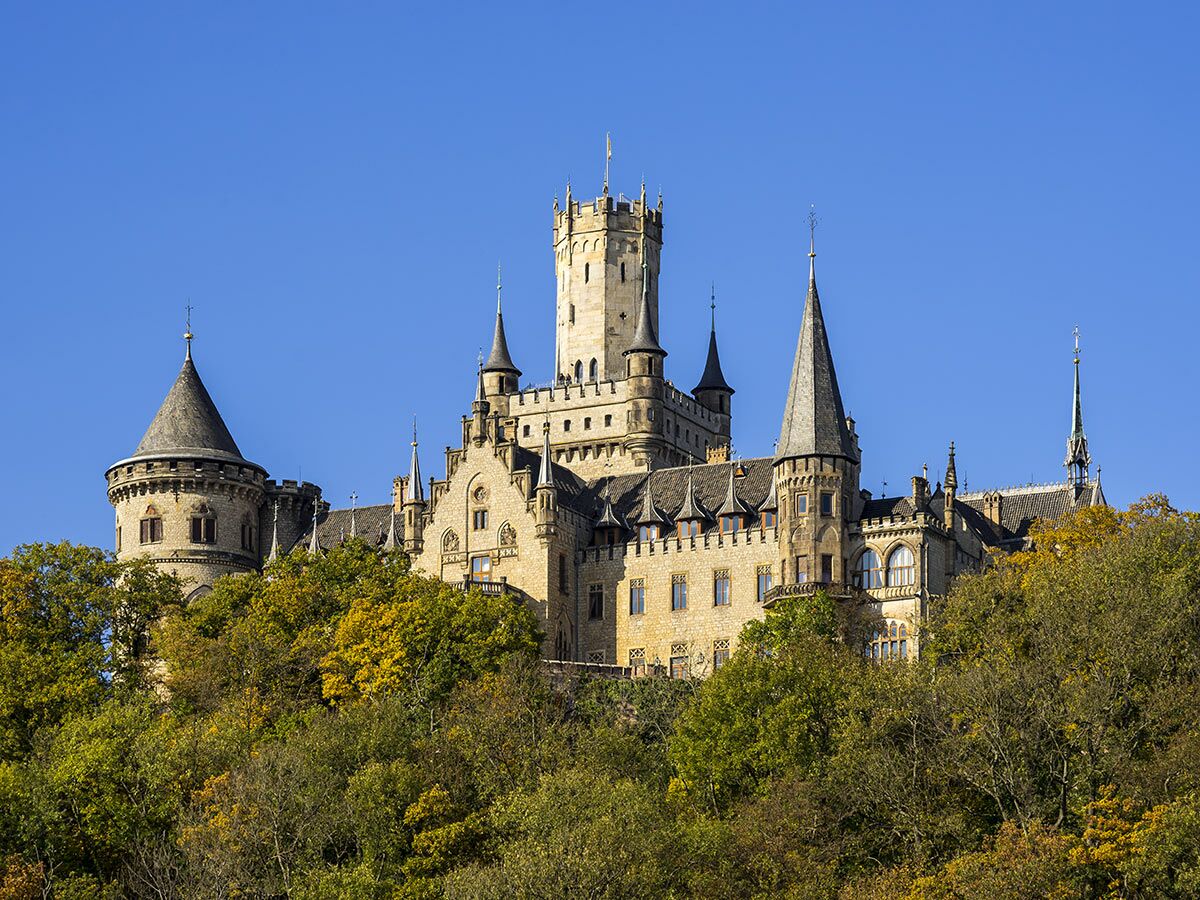 Das majestätische Marienburg Schloss thront auf einem bewaldeten Hügel, umgeben von herbstlichen Bäumen unter blauem Himmel.