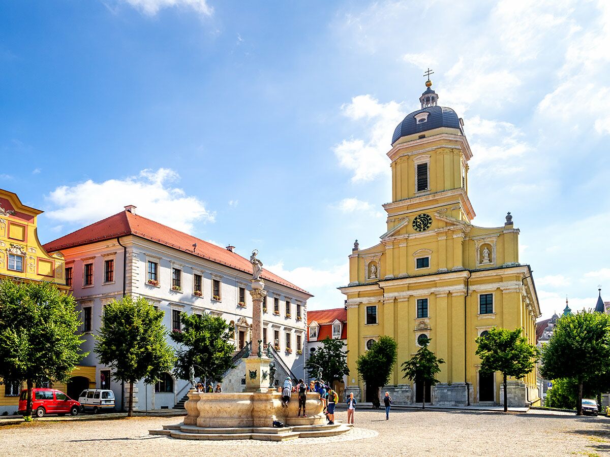 Gelbe Barockkirche und Mariensäule Brunnen auf historischem Marktplatz in Dillingen an der Donau unter blauem Himmel mit Menschen.
