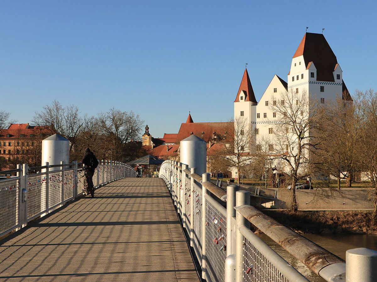 Eine Person geht über eine Fußgängerbrücke mit Liebesschlössern in Ingolstadt, mit dem weißen Neuen Schloss im Hintergrund.