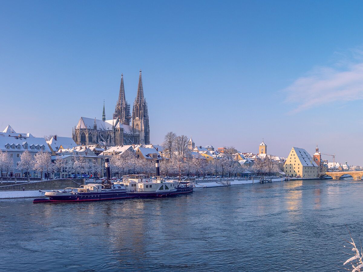 Schneebedeckte Altstadt von Regensburg im Winter: Blick über die Donau auf den Dom, historische Gebäude und Boote.