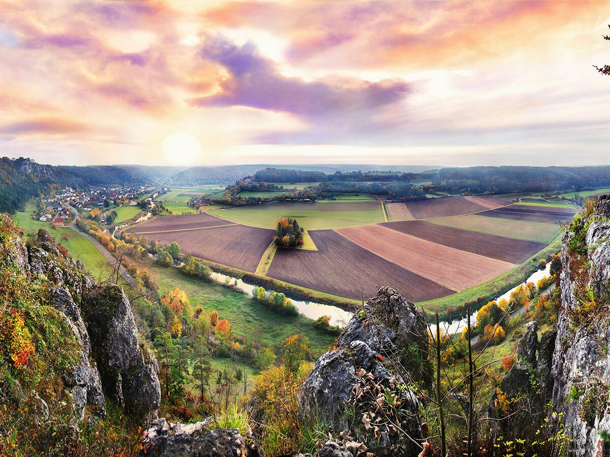 Panoramaaufnahme einer herbstlichen Flusslandschaft mit Dorf, Feldern und bunten Bäumen bei Sonnenuntergang aus Felsenblick.