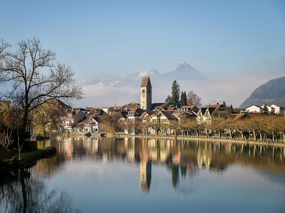 Malerische Alpenstadt mit traditionellen Häusern, Kirchturm und schneebedeckten Bergen, gespiegelt in einem ruhigen See oder Fluss.