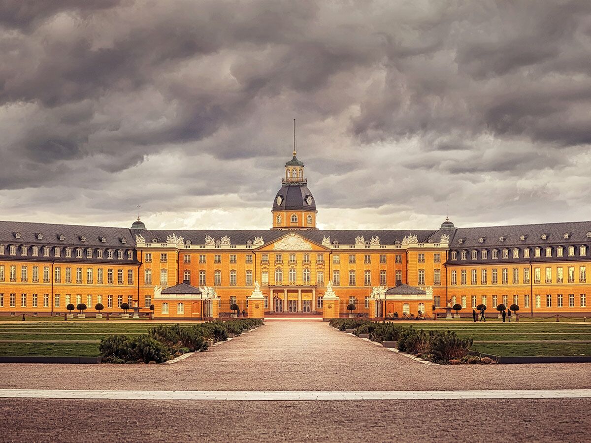 Majestätisches Karlsruher Schloss mit gelber Fassade, dunklen Dächern und zentralem Turm unter dramatischem Wolkenhimmel.