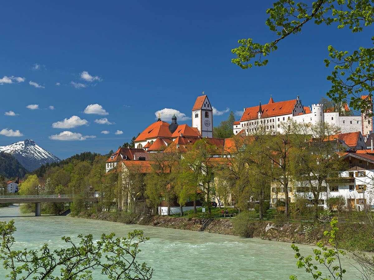 Panoramablick auf die historische Stadt Füssen mit Hohem Schloss, St. Mang Basilika, Lech Fluss und schneebedeckten Alpen im Frühling.