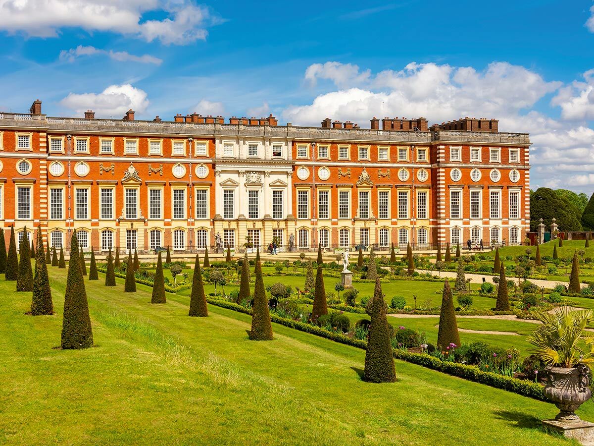 Hampton Court Palast und seine formalen Gärten mit Kegel-Formschnittbäumen unter blauem Himmel an einem Sommertag.
