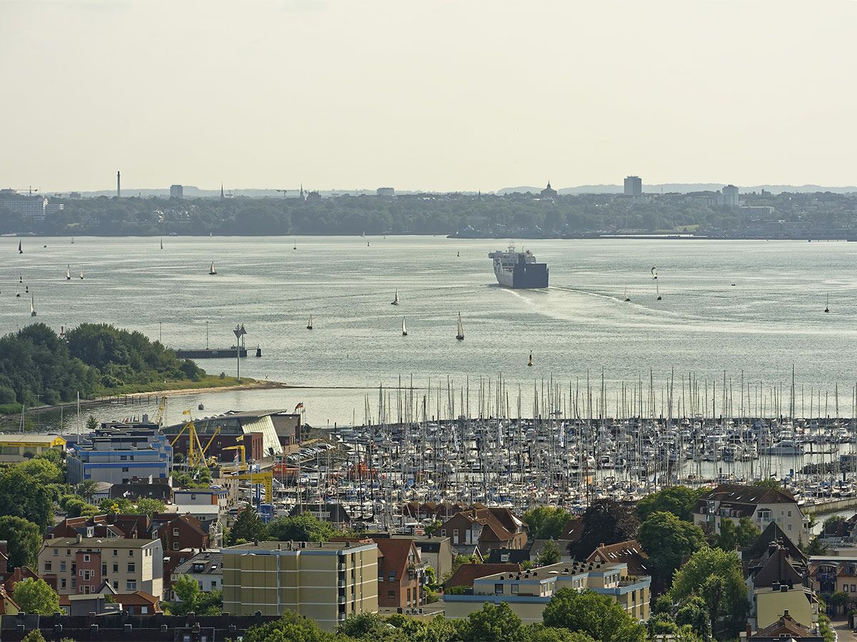 Panoramablick auf eine Hafenstadt mit belebter Marina voller Segelboote, einem Frachtschiff auf dem Wasser und Gebäuden im Vordergrund.
