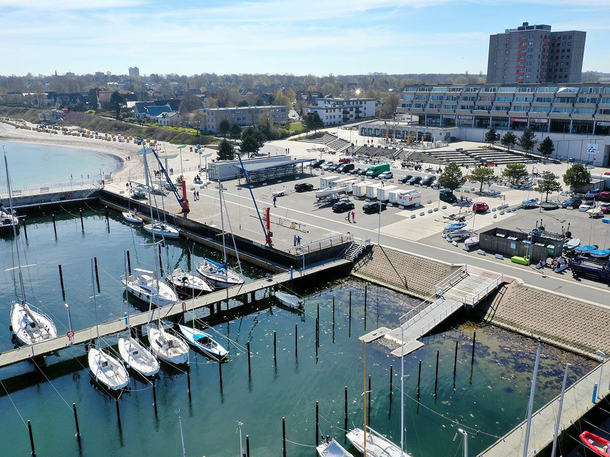Luftaufnahme eines belebten Yachthafens mit zahlreichen Segelbooten, angrenzendem Strand und Stadtgebäuden bei Sonnenschein.