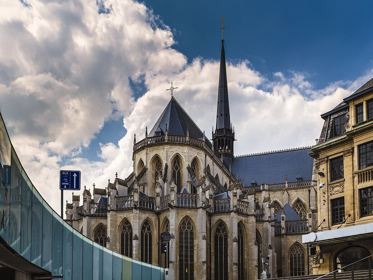 Große gotische Kathedrale mit hohem Turm und Buntglasfenstern, neben einem modernen geschwungenen Glasgebäude. Blauer Himmel mit Wolken.