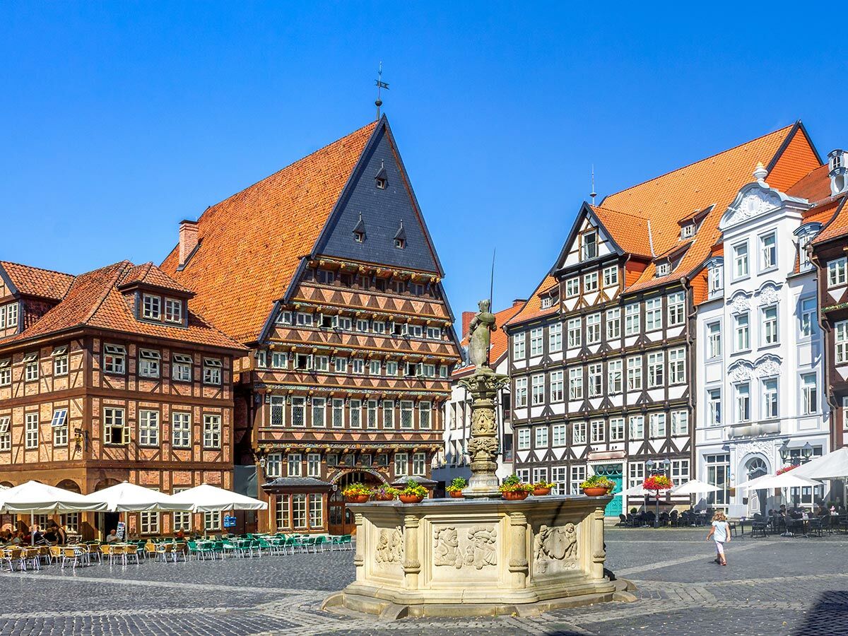 Historischer Marktplatz in Hildesheim mit prächtigen Fachwerkhäusern, zentralem Brunnen und belebten Straßencafés unter blauem Himmel.