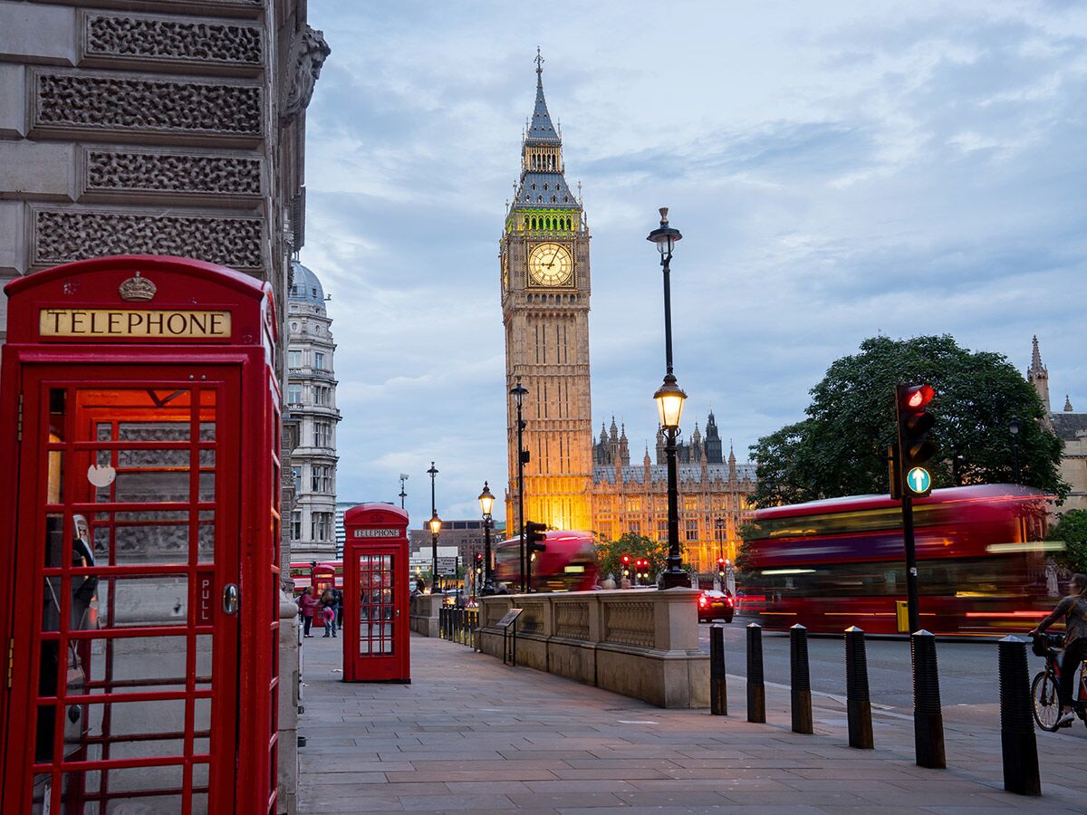 Abend in London: Big Ben, Houses of Parliament, rote Telefonzellen und roter Doppeldeckerbus mit Bewegungsunschärfe auf Straße.