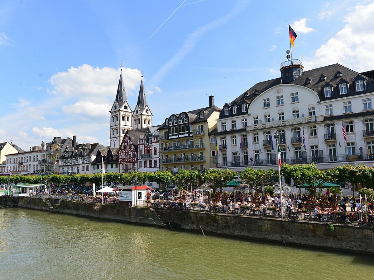 Blick auf die belebte Rheinpromenade von Bacharach mit Terrassencafés, Fachwerkhäusern, der St. Peter Kirche und dem Hotel Bellevue.