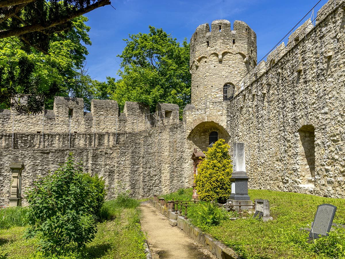 Historische Steinmauer einer Burg mit rundem Turm, Zinnen, einem alten Friedhof und Bäumen unter blauem Himmel.