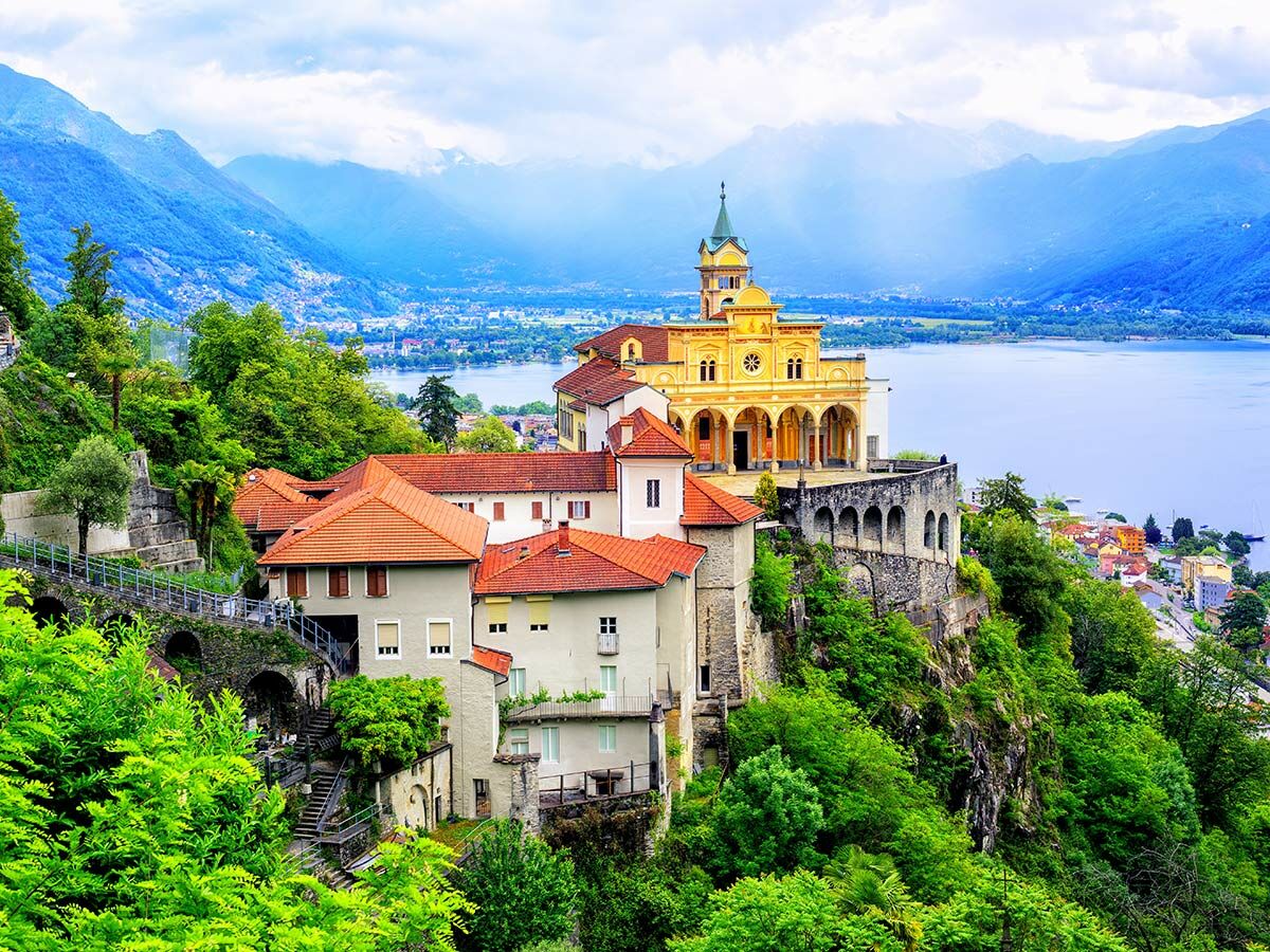 Die goldene Wallfahrtskirche Madonna del Sasso auf einem grünen Hügel mit Häusern und einem See im Hintergrund, umgeben von Bergen unter blauem Himmel.